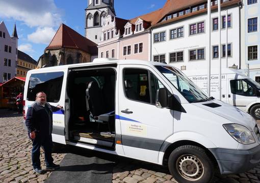 Mann um die 60 an weißen Minibus gelehnt lächelnd vor Stadtkulisse